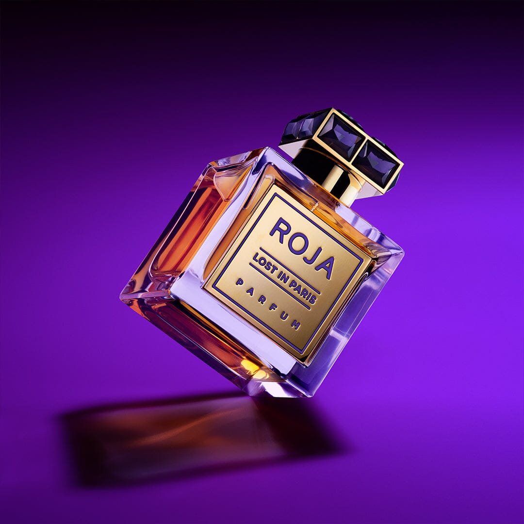 A luxurious glass bottle of Roja Parfums Lost in Paris, featuring gourmand accords, a golden label, and dark cap, is displayed against a vibrant purple background with light reflections and shadows.
