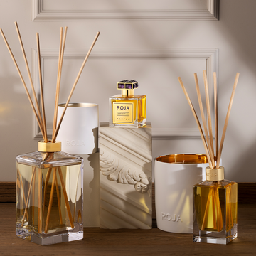 A selection of ROJA London luxury fragrance items, featuring a Lost in Paris diffuser with gourmand notes, two golden-yellow reed diffusers, a gold-capped perfume bottle, and decorative containers displayed on wood.