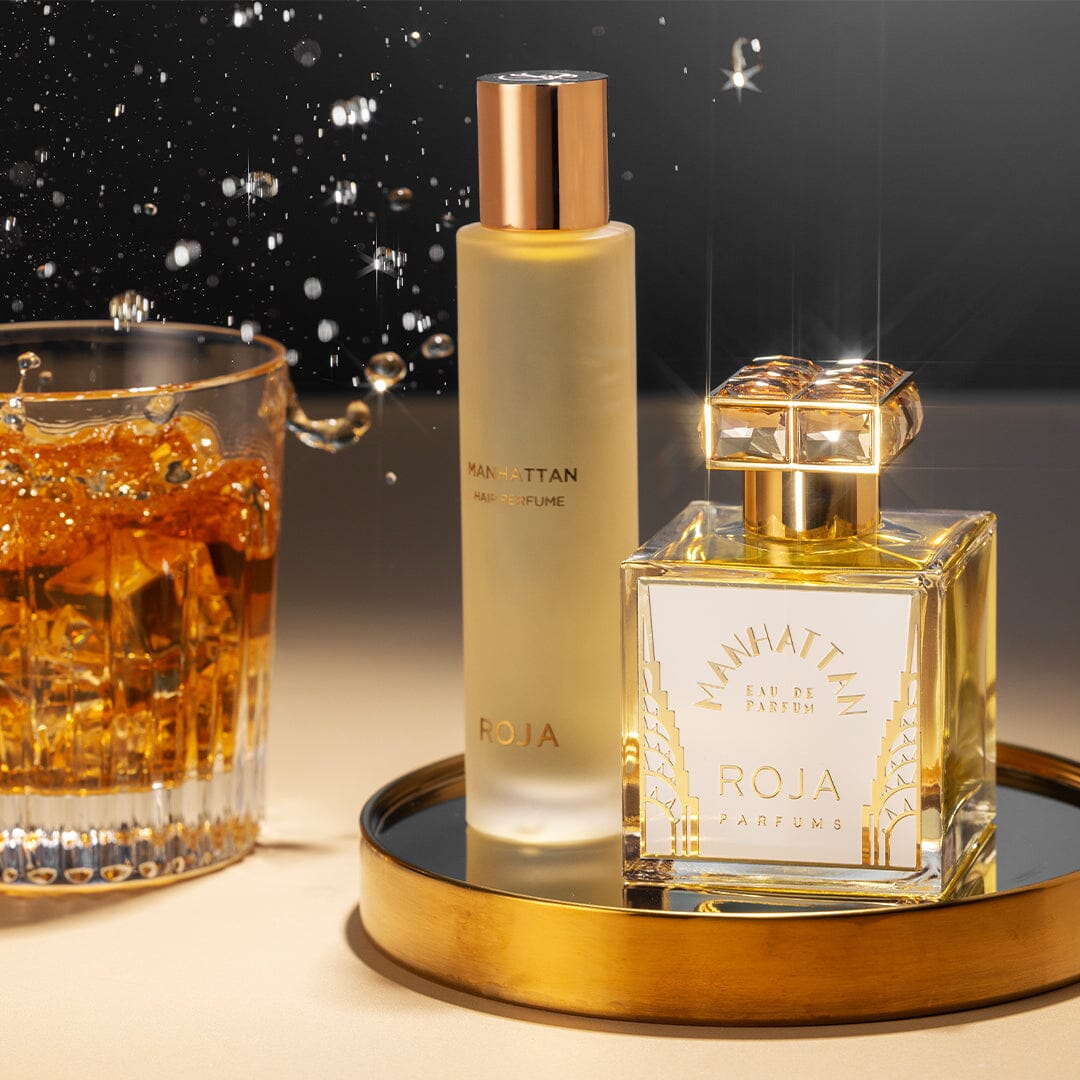 Two bottles of Manhattan Hair Mist by Roja Parfums—one tall and frosted, the other square with a gold cap—sit on a gold tray beside a whiskey glass, evoking cedarwood tobacco notes in an elegant, glamorous setting.