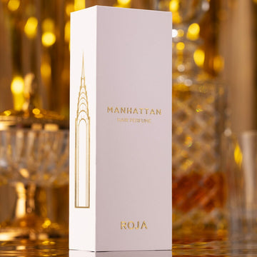 A tall, white rectangular box of Manhattan Hair Perfume by Roja stands upright. The box features gold lettering and an elegant gold design, with a blurred, luxurious background of glass and gold décor.