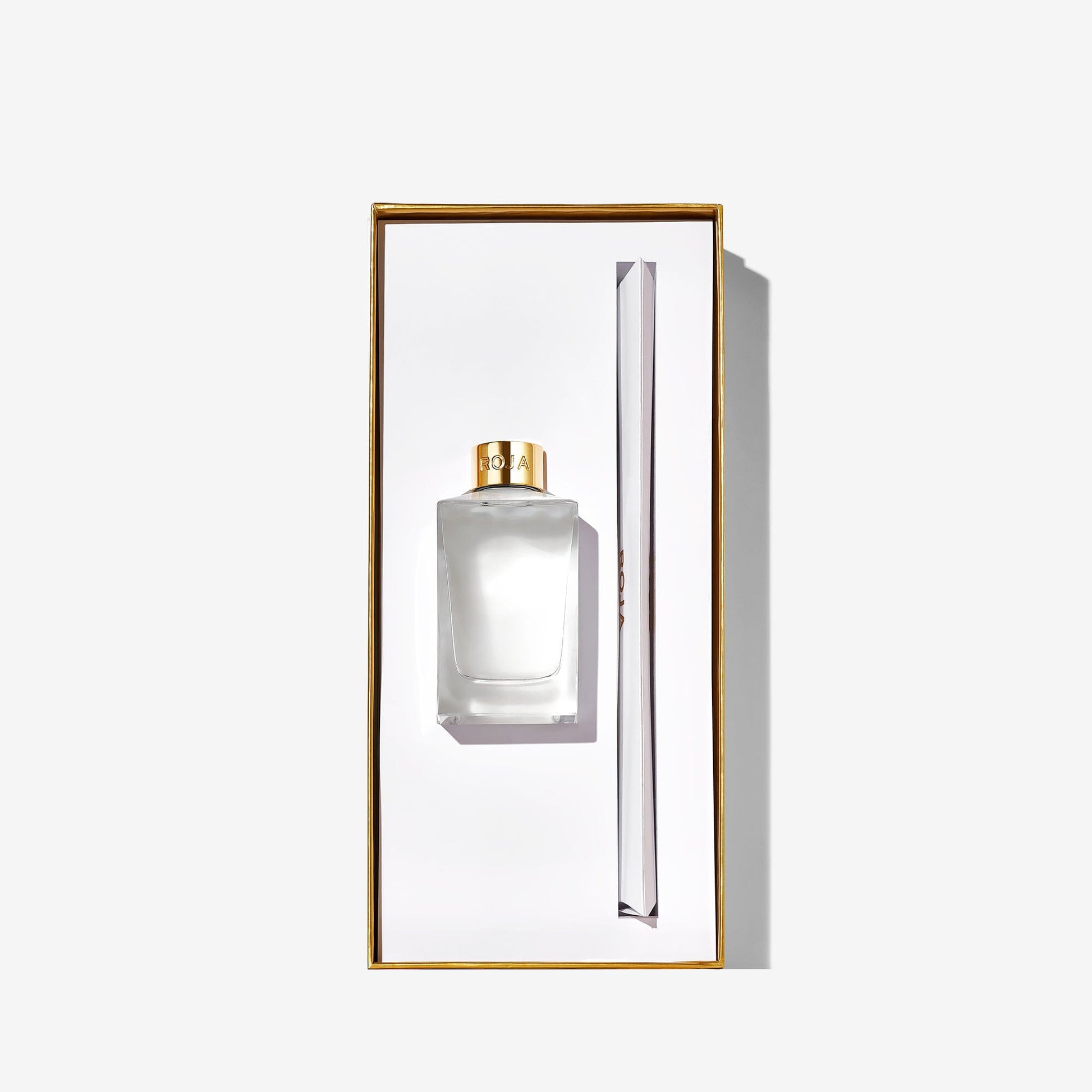 A rectangular box contains a clear Roja Parfums New York perfume bottle with a gold cap and slim applicator, set against a white background with gold edging—an elegant design inspired by Central Park.