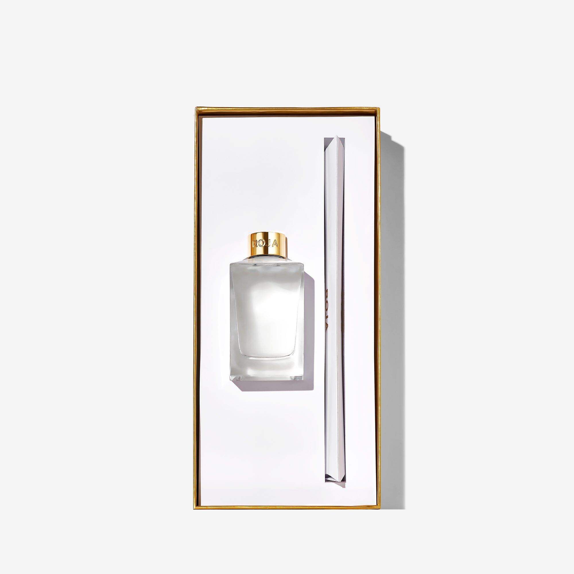 A rectangular box contains a clear Roja Parfums New York perfume bottle with a gold cap and slim applicator, set against a white background with gold edging—an elegant design inspired by Central Park.