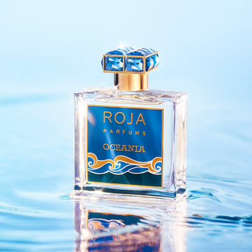 A perfume bottle labeled The Paradise Duet Gift Set by Roja Parfums rests on a reflective water surface. Adorned with a blue-and-gold design, wave patterns, and jeweled cap, the scene shimmers in soft blues and sparkles, capturing the serene allure reminiscent of Isola Blu.
