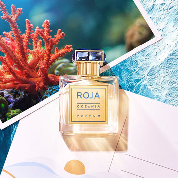 A bottle of Roja Oceania Parfum rests on top of a postcard and a photo of coral underwater, evoking a fresh and aquatic theme. The perfume bottle has a gold cap and a blue and gold label.