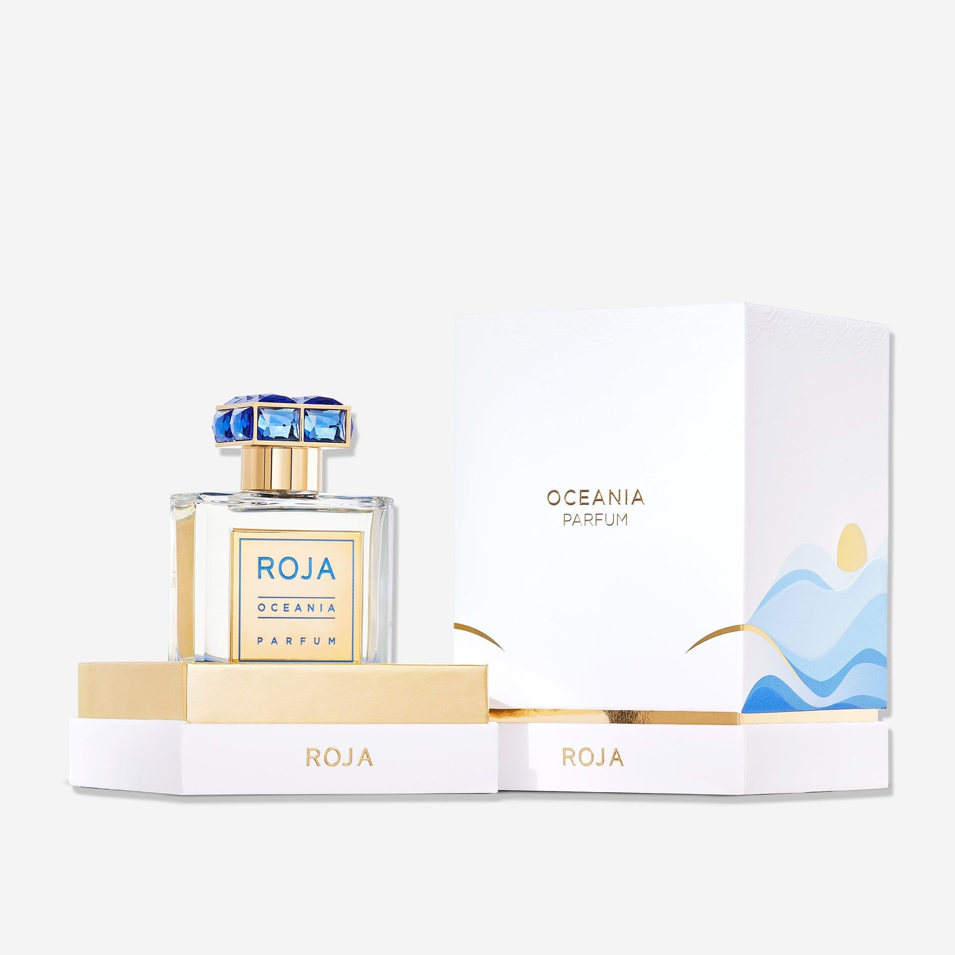 A bottle of Roja Parfums Oceania with a blue and gold cap sits beside its white and gold box, adorned with blue wave designs and gold lettering—a stylish tribute to the Isola Collection.