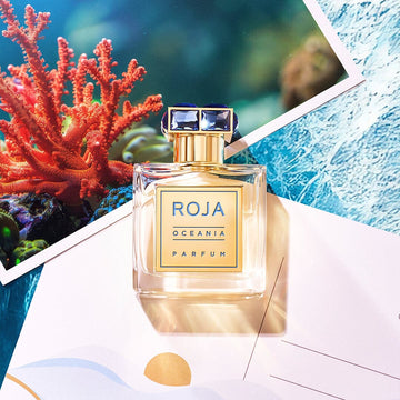 A bottle of Roja Oceania Parfum sits on top of two colorful postcards, one featuring a coral and underwater scene, with sunlight illuminating the bottle and creating reflections.