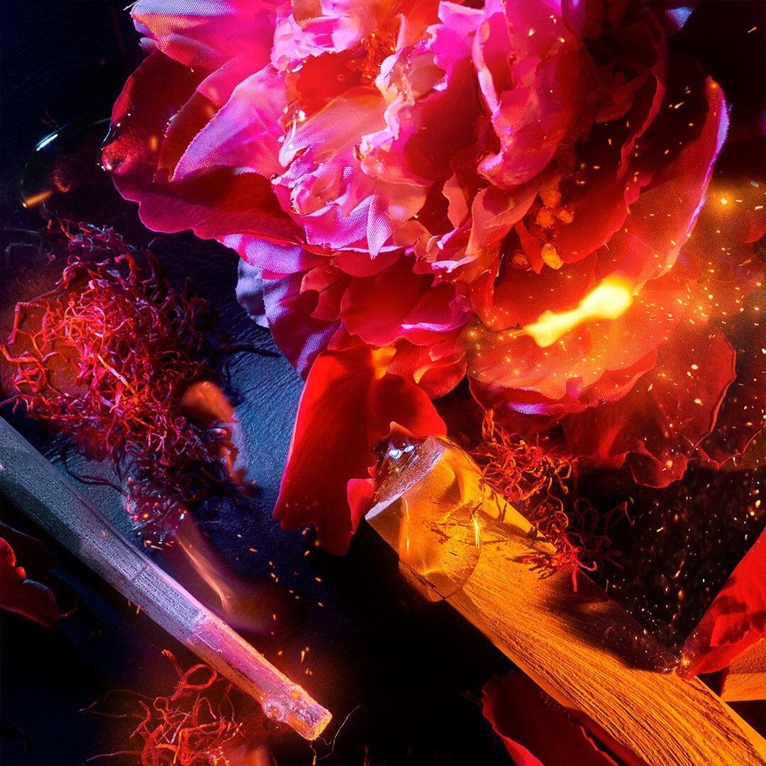 Vivid close-up of a pink peony flower surrounded by scattered saffron threads, a glass vial, and glowing sparks, all set against a dark background.