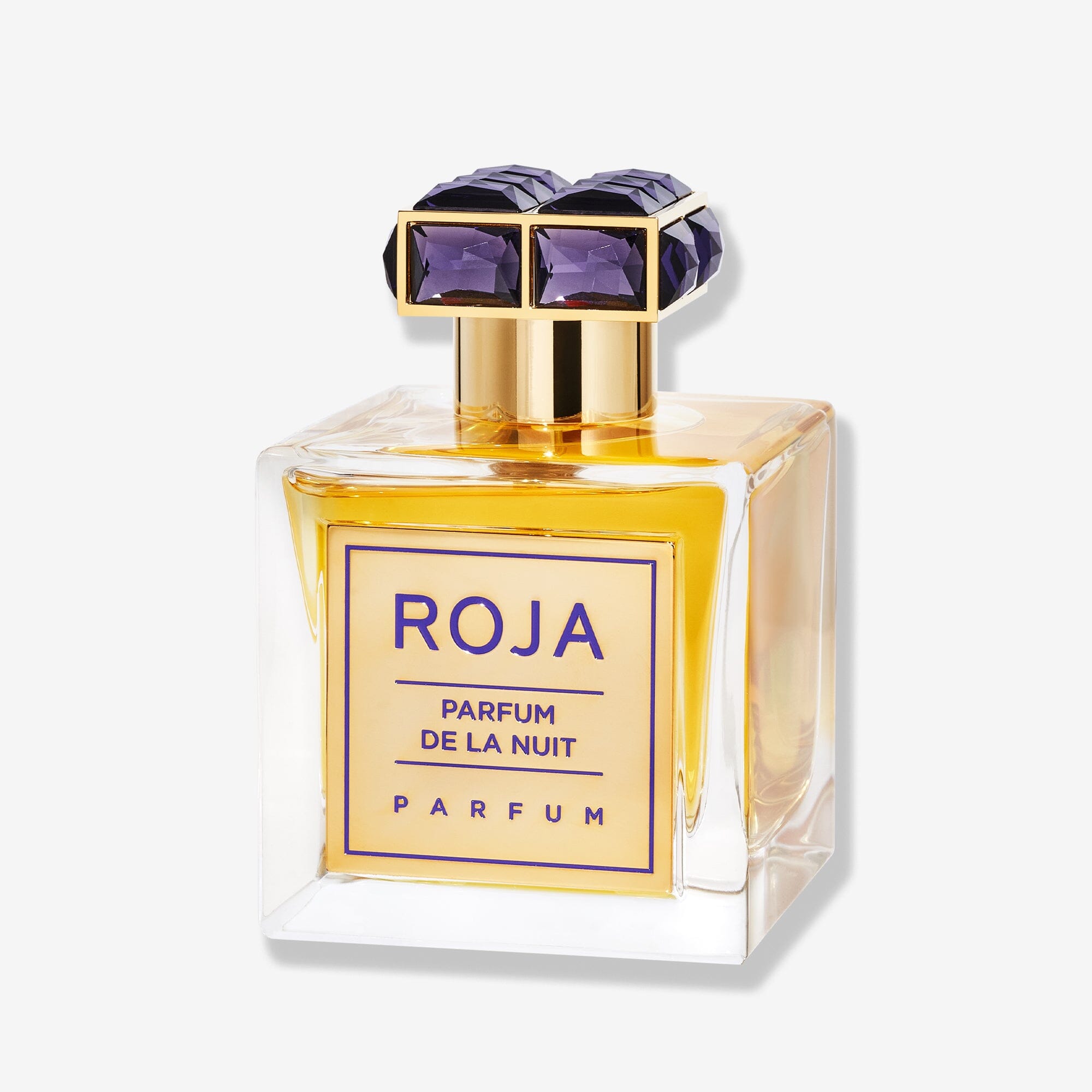 A square glass bottle of Roja Parfums Parfum De La Nuit, an amber-hued scent with gold accents and a cap adorned with purple gemstones. The label features blue text on gold, highlighting this sensual fragrance.