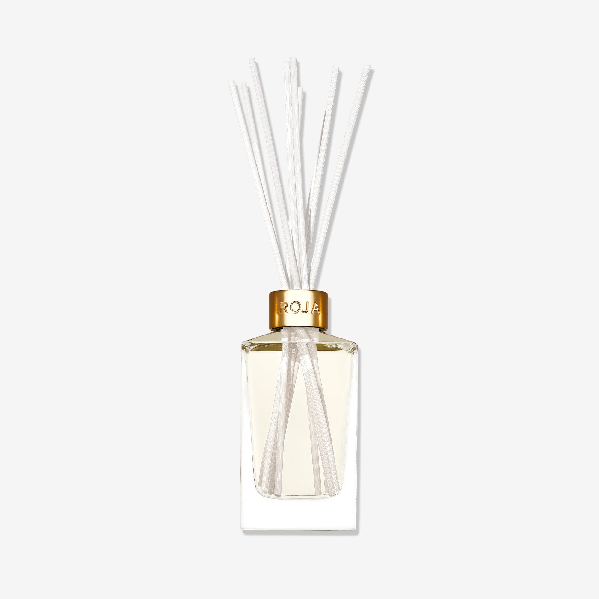 The Tea Time In The Conservatory reed diffuser by Roja Parfums features a clear glass vessel with a gold cap, light-hued liquid, and white reeds fanning out, evoking the subtle scent of Jasmine Tea against a plain white background.
