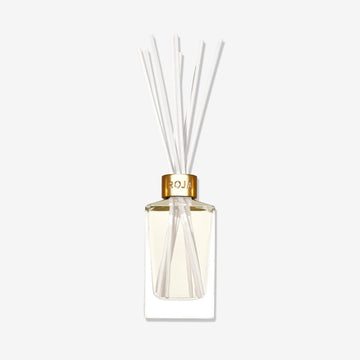 The Tea Time In The Conservatory reed diffuser by Roja Parfums features a clear glass vessel with a gold cap, light-hued liquid, and white reeds fanning out, evoking the subtle scent of Jasmine Tea against a plain white background.