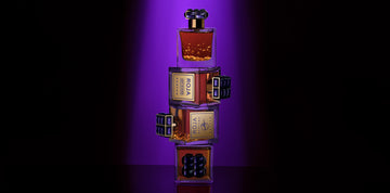 Four perfume bottles with gold labels and dark caps are stacked vertically against a purple-lit background, creating a dramatic and elegant display.