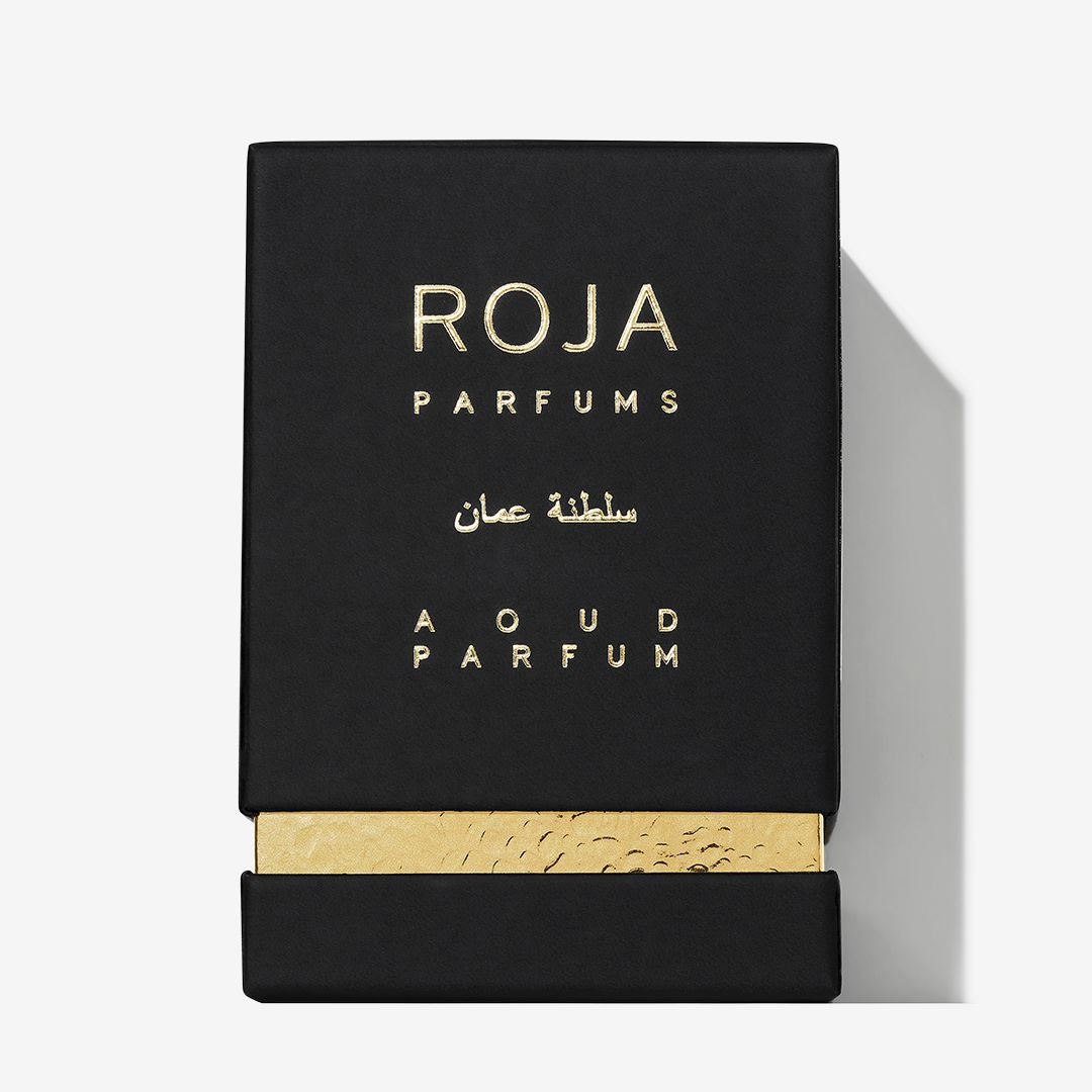 A black rectangular luxury fragrance box for Roja Parfums Qatar, featuring gold-embossed branding and Arabic script. A gold textured band near the bottom adds a touch of classic Middle Eastern perfume elegance.