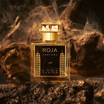 A luxurious gold perfume bottle labeled Roja Parfums Aoud Parfum sits on dark textured wood, surrounded by smoky swirls, against a dramatic, shadowy background.
