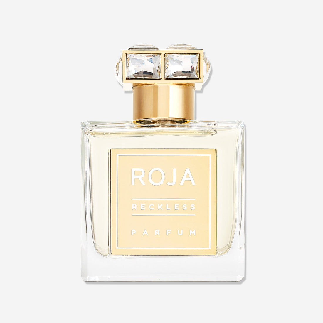 A clear glass bottle of Roja Parfums Reckless Pour Femme with a gold label and jeweled cap is displayed against a white background. This summery scent offers sparkling pink pepper notes for an elegant, refreshing feel.