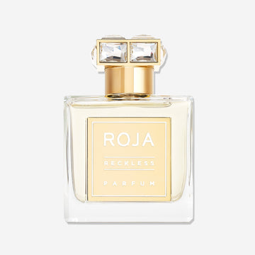 A clear glass bottle of Roja Parfums Reckless Pour Femme with a gold label and jeweled cap is displayed against a white background. This summery scent offers sparkling pink pepper notes for an elegant, refreshing feel.