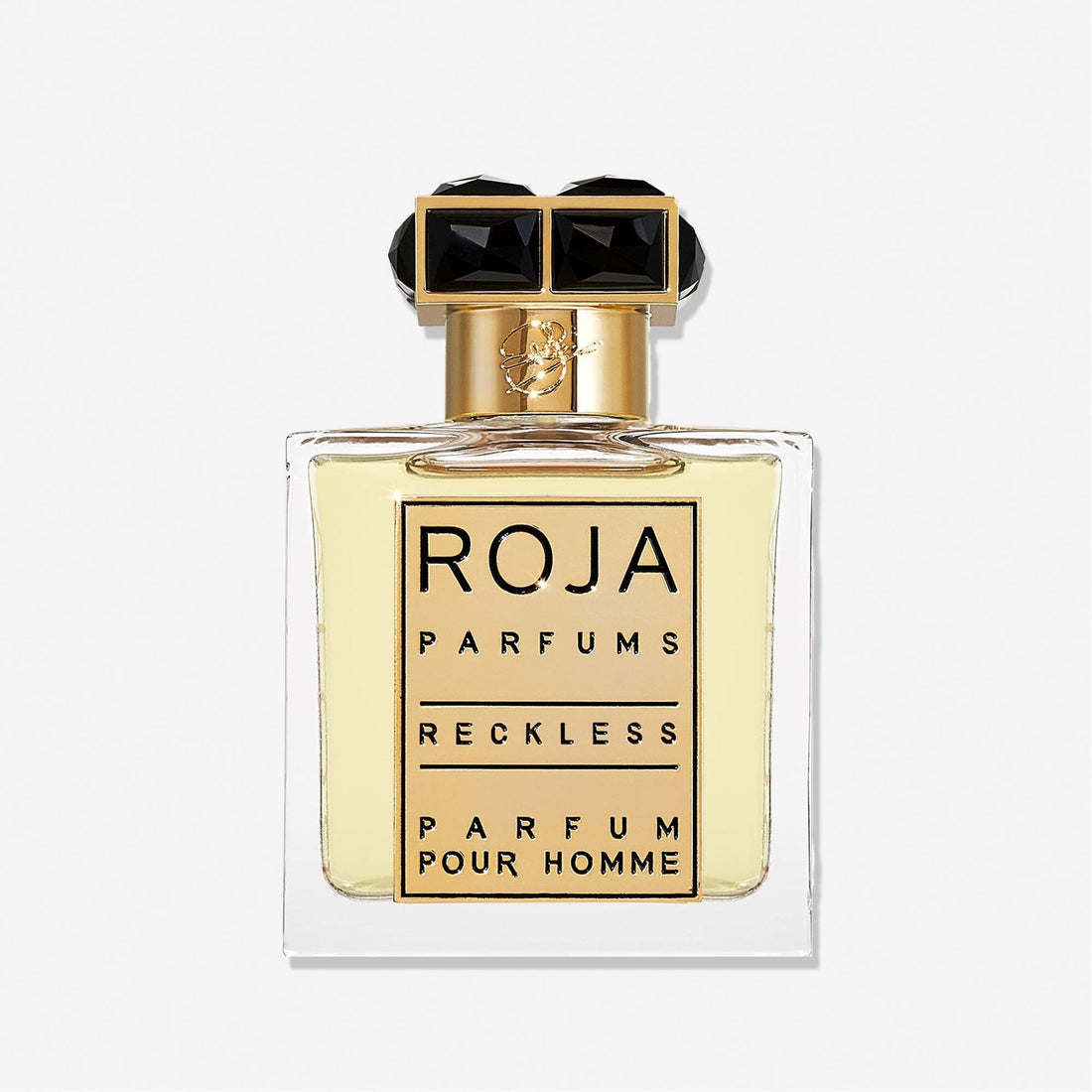 A clear glass bottle of Roja Parfums Reckless Pour Homme features a gold and black cap and holds light yellow liquid. The label shows the brand and product name in bold black letters—a masculine scent with spicy chypré notes.