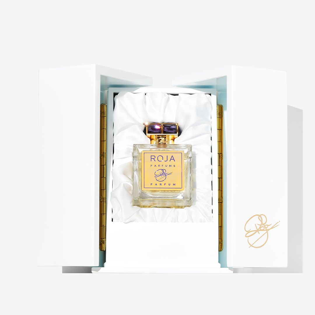 An elegant bottle of ROJA by Roja Parfums stands upright in a white satin-lined box with gold accents and a signature, creating an exquisite presentation for this renowned luxury fragrance.