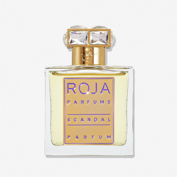A clear glass bottle of Roja Parfums Scandal Pour Femme features a gold label with purple text and a gold cap set with two large, square, clear gemstones—an elegant nod to jasmine’s timeless allure.