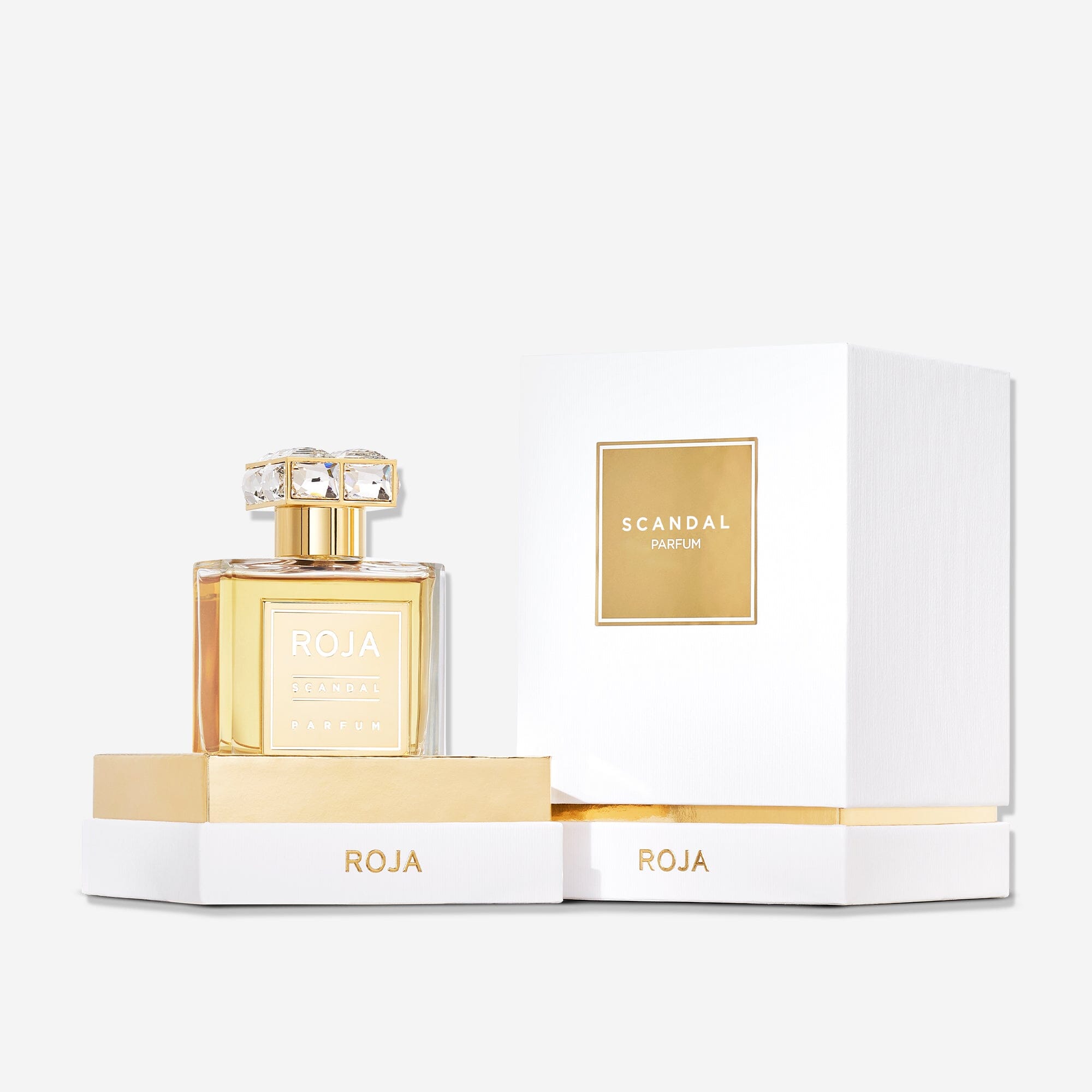 A bottle of Roja Parfums Scandal Pour Femme with a gold label stands beside its open white and gold box on a white background—an exquisite fragrance with sandalwood notes.