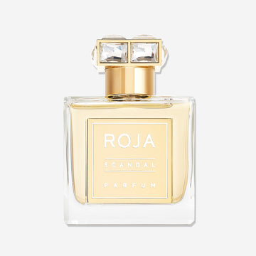 A clear glass perfume bottle with gold accents and a square gold label reading Roja Parfums Scandal Pour Femme. The gold cap features two large, clear rectangular gems, evoking the allure of narcotic white florals like jasmine and tuberose.