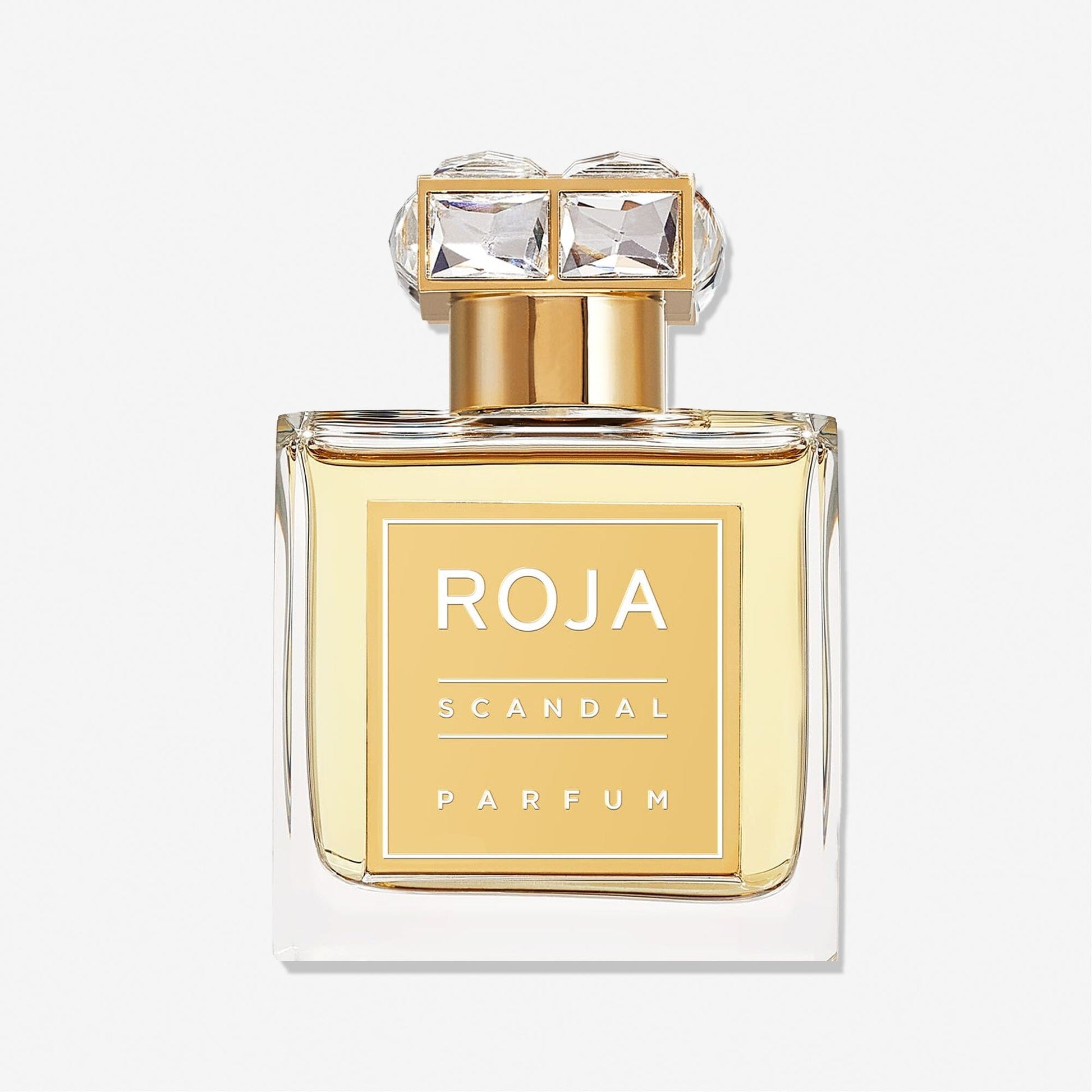 A clear glass bottle of Roja Parfums Scandal Pour Femme, featuring a gold cap with two large square crystals, stands against a plain white background and evokes the essence of scandal with hints of Sandalwood Musk.