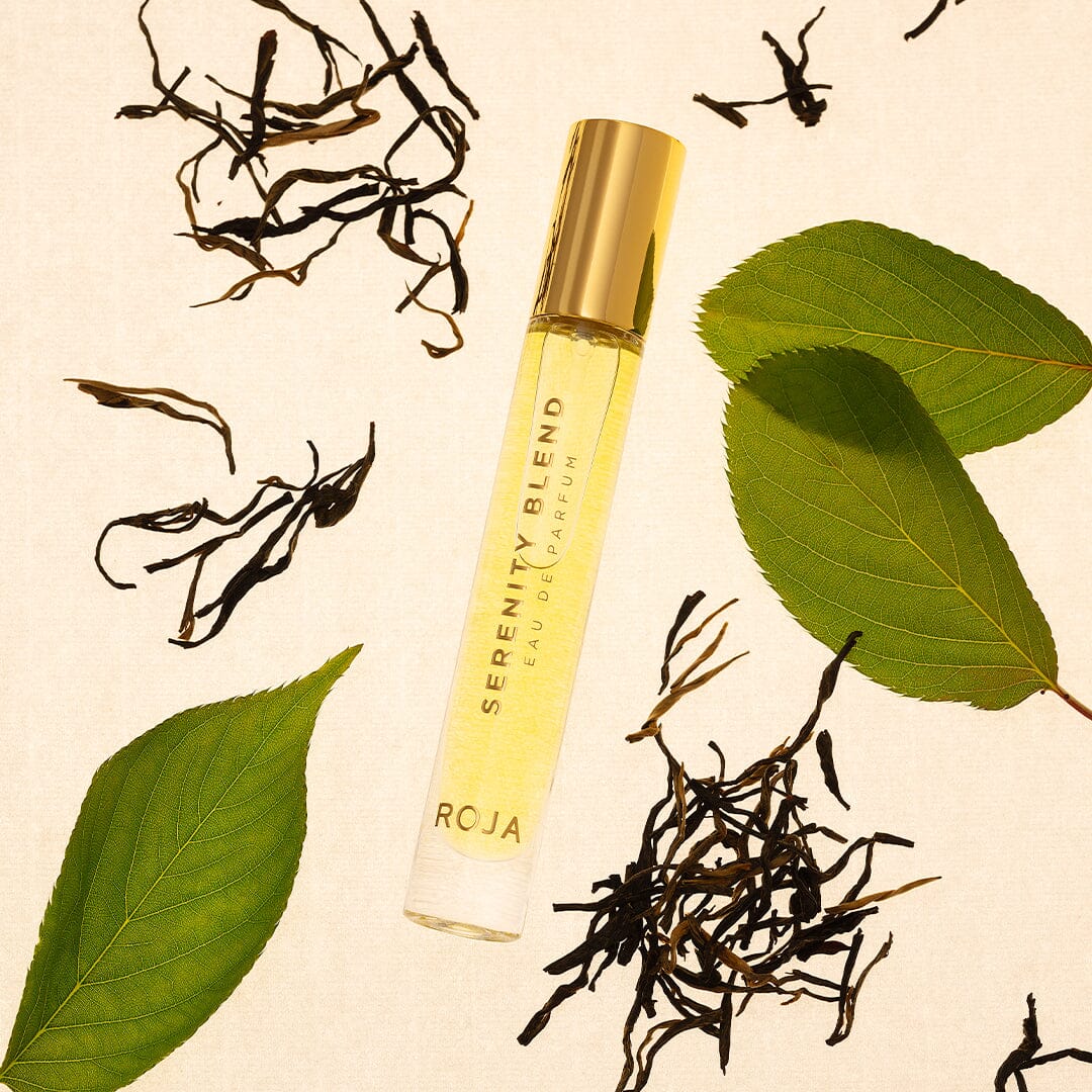 A 10ml Serenity Blend Eau De Parfum vial by Roja Parfums with a gold cap rests on beige, surrounded by green leaves and dark tea leaves.