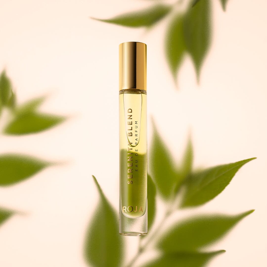 A 10ml bottle of Serenity Blend Eau De Parfum by Roja Parfums with a gold cap stands upright among green leaves on a soft beige background, featuring the brand name at the base.