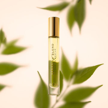 A 10ml bottle of Serenity Blend Eau De Parfum by Roja Parfums with a gold cap stands upright among green leaves on a soft beige background, featuring the brand name at the base.