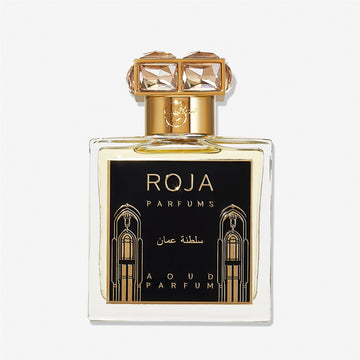 A clear perfume bottle labeled Roja Parfums Sultanate of Oman features gold accents, a black and gold label with Arabic designs, and a crystal-embellished gold cap—evoking luxurious Aoud and smoky frankincense.