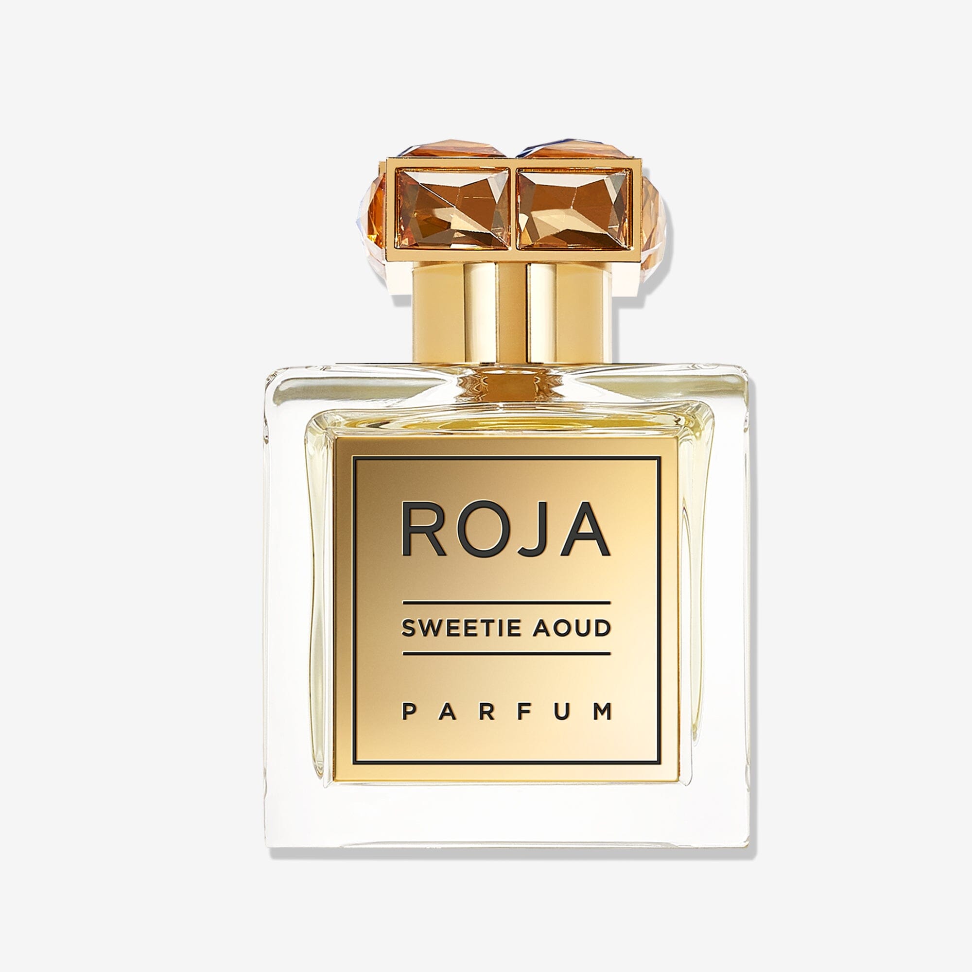 A clear glass bottle of Roja Parfums Sweetie Aoud, with a gold label and cap adorned by two square amber gemstones, stands against a plain white background.