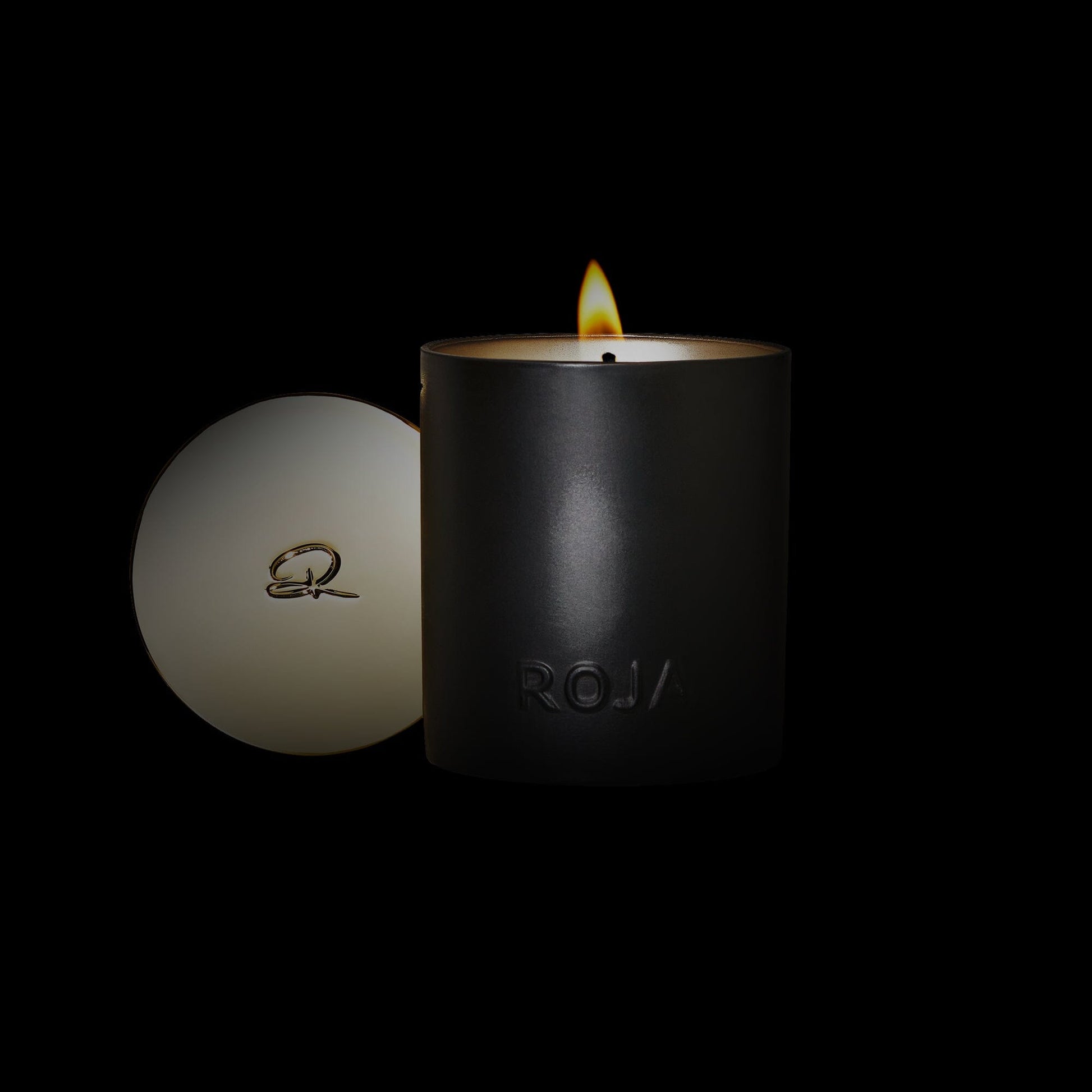 A black Amber Aoud candle by Roja Parfums burns softly beside its round lid with a cursive R, set against a dark background, evoking olfactive majesty.