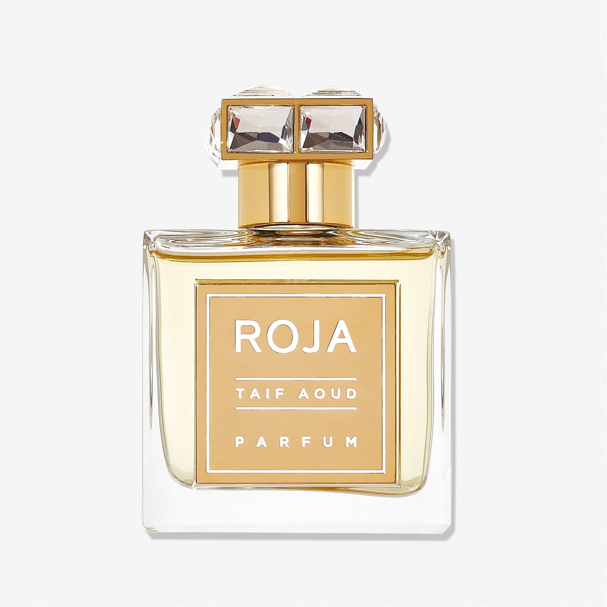 A clear glass bottle of Roja Parfums Taif Aoud, featuring a gold label and square jeweled cap with light golden liquid, captures the essence of Arabian rose, displayed against a plain white background.