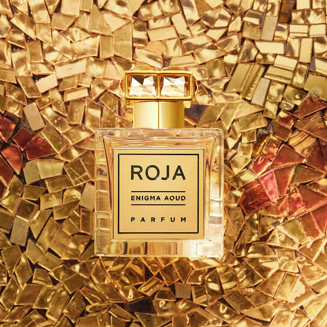A square glass bottle of Roja Enigma Aoud Parfum with a gold cap is placed on a mosaic background of gold and metallic tiles, creating a luxurious and elegant look.