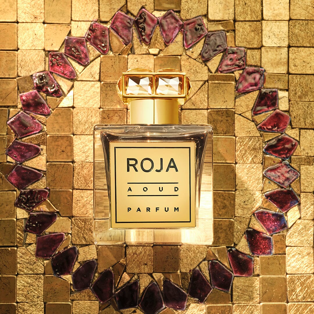 A bottle of Roja Aoud Parfum sits on a gold mosaic tile surface, framed by a circular pattern of purple and gold tiles.