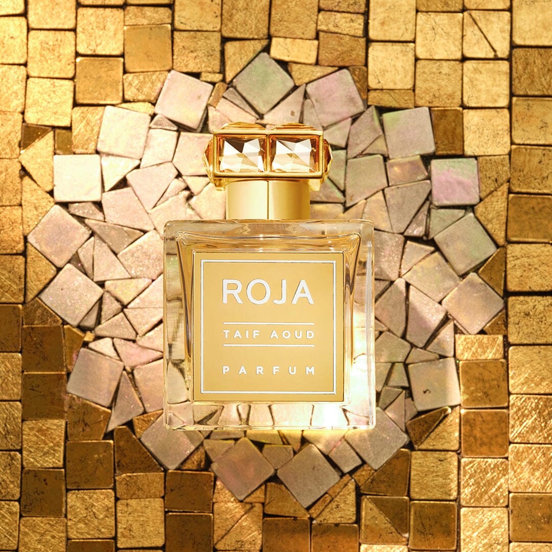 A bottle of Roja Taif Aoud Parfum with a gold cap is placed on a mosaic background of shiny gold and iridescent tiles, creating a luxurious and elegant atmosphere.