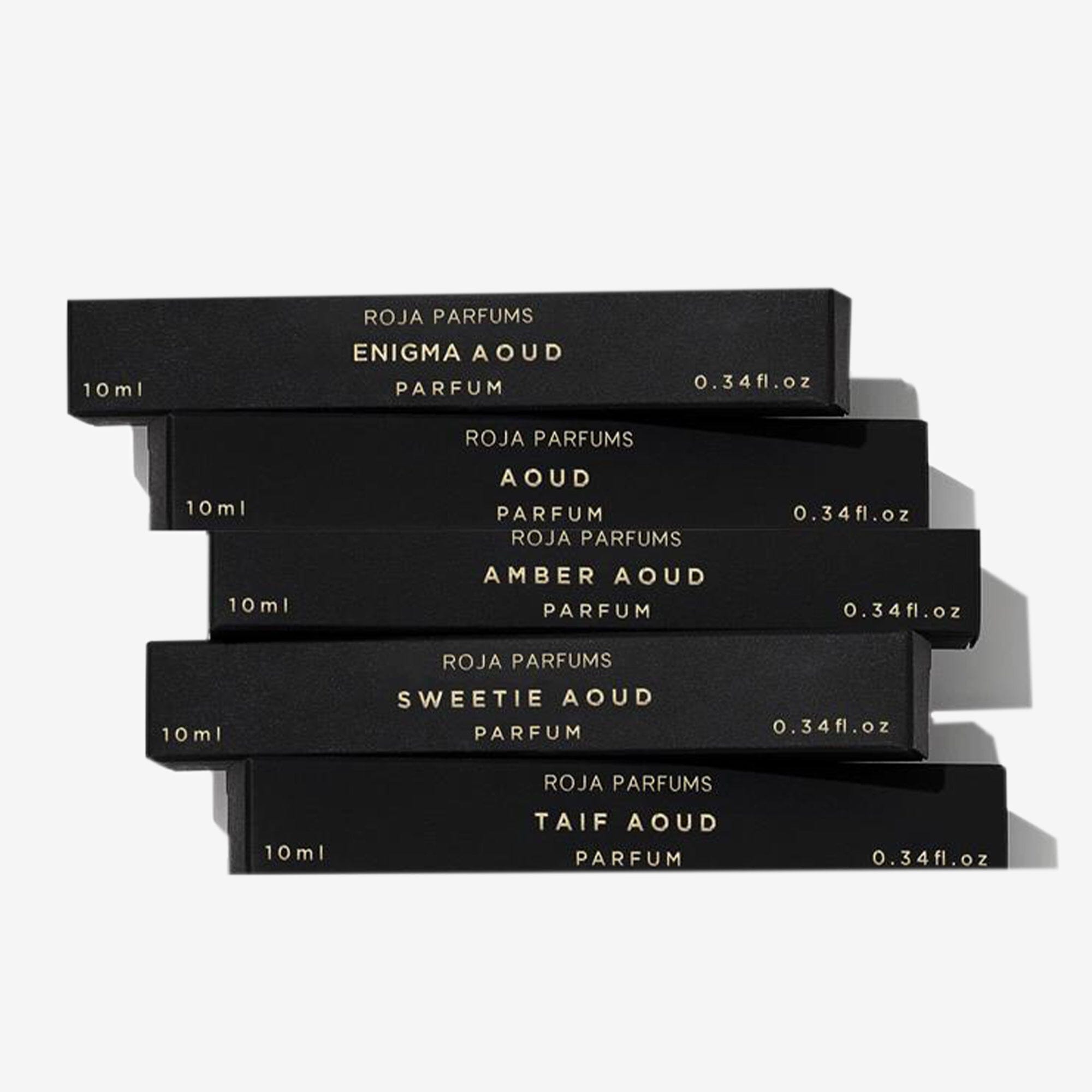 Five sleek black perfume boxes with gold lettering are stacked askew. The Aoud Travel Collection by Roja Parfums includes 10ml scents: Enigma Aoud, Aoud, Amber Aoud, Sweetie Aoud, and Taif Aoud.