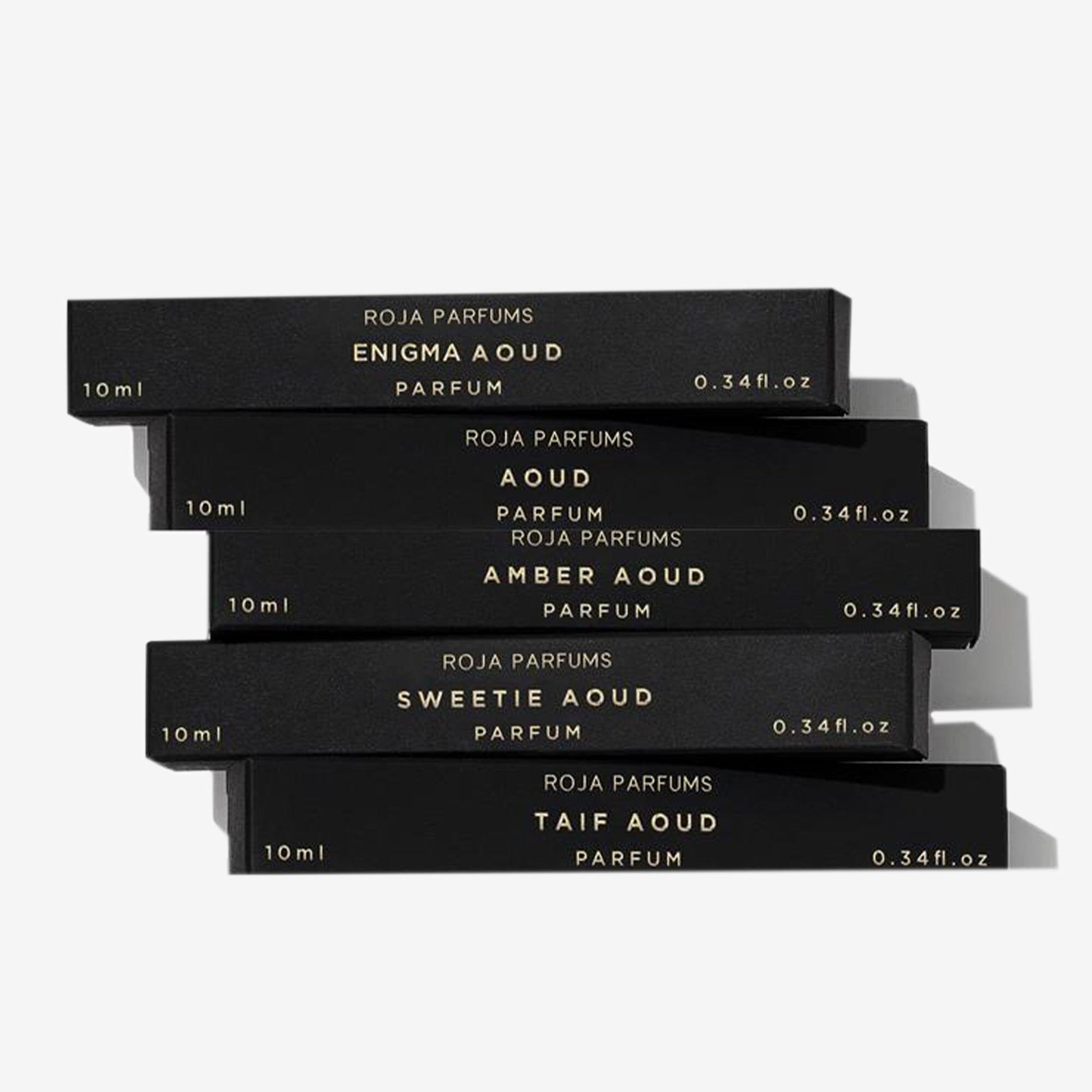 Five sleek black perfume boxes with gold lettering are stacked askew. The Aoud Travel Collection by Roja Parfums includes 10ml scents: Enigma Aoud, Aoud, Amber Aoud, Sweetie Aoud, and Taif Aoud.