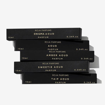 Five sleek black perfume boxes with gold lettering are stacked askew. The Aoud Travel Collection by Roja Parfums includes 10ml scents: Enigma Aoud, Aoud, Amber Aoud, Sweetie Aoud, and Taif Aoud.