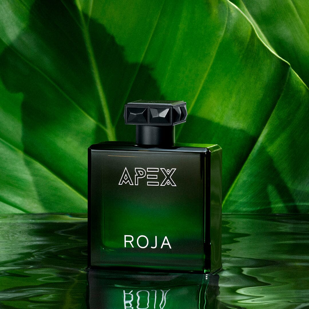 A green glass bottle from The Apex Gift Set by Roja Parfums sits on a reflective surface amid lush tropical leaves. Featuring a geometric black cap and bold white lettering, it perfectly represents the Art Of Layering in fragrance.