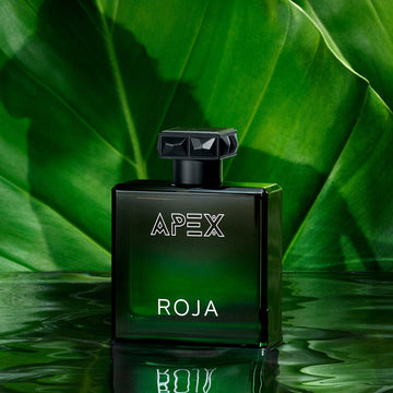 A green glass bottle from The Apex Gift Set by Roja Parfums sits on a reflective surface amid lush tropical leaves. Featuring a geometric black cap and bold white lettering, it perfectly represents the Art Of Layering in fragrance.