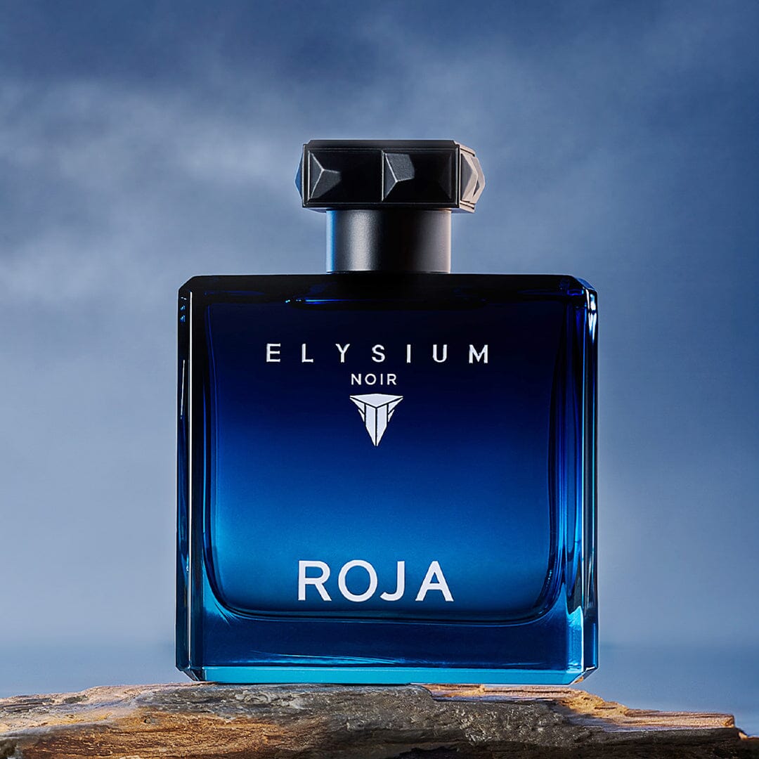 A blue glass bottle from The Elysium Trio by ROJA London, featuring a black geometric cap on a wooden surface with a cloudy blue backdrop—ideal for elevating your fragrance collection and mastering the Art Of Layering.
