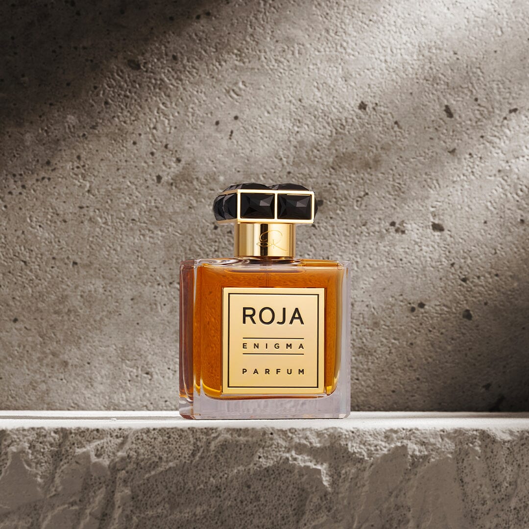 A rectangular Roja Parfums Enigma Pour Homme glass perfume bottle filled with amber cognac cologne and topped with a faceted black cap, sitting on a stone ledge.