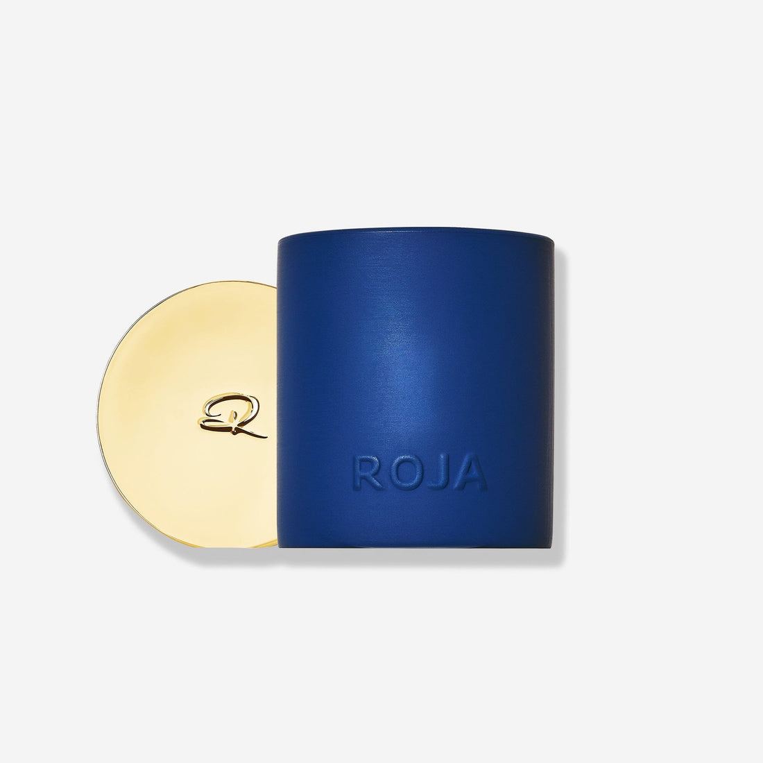 A matte blue cylindrical container embossed with "ROJA" sits beside a round gold lid with a decorative "R," showcasing Roja Parfums’ The Essence Of Christmas candle—a festive home fragrance—set against a plain white background.