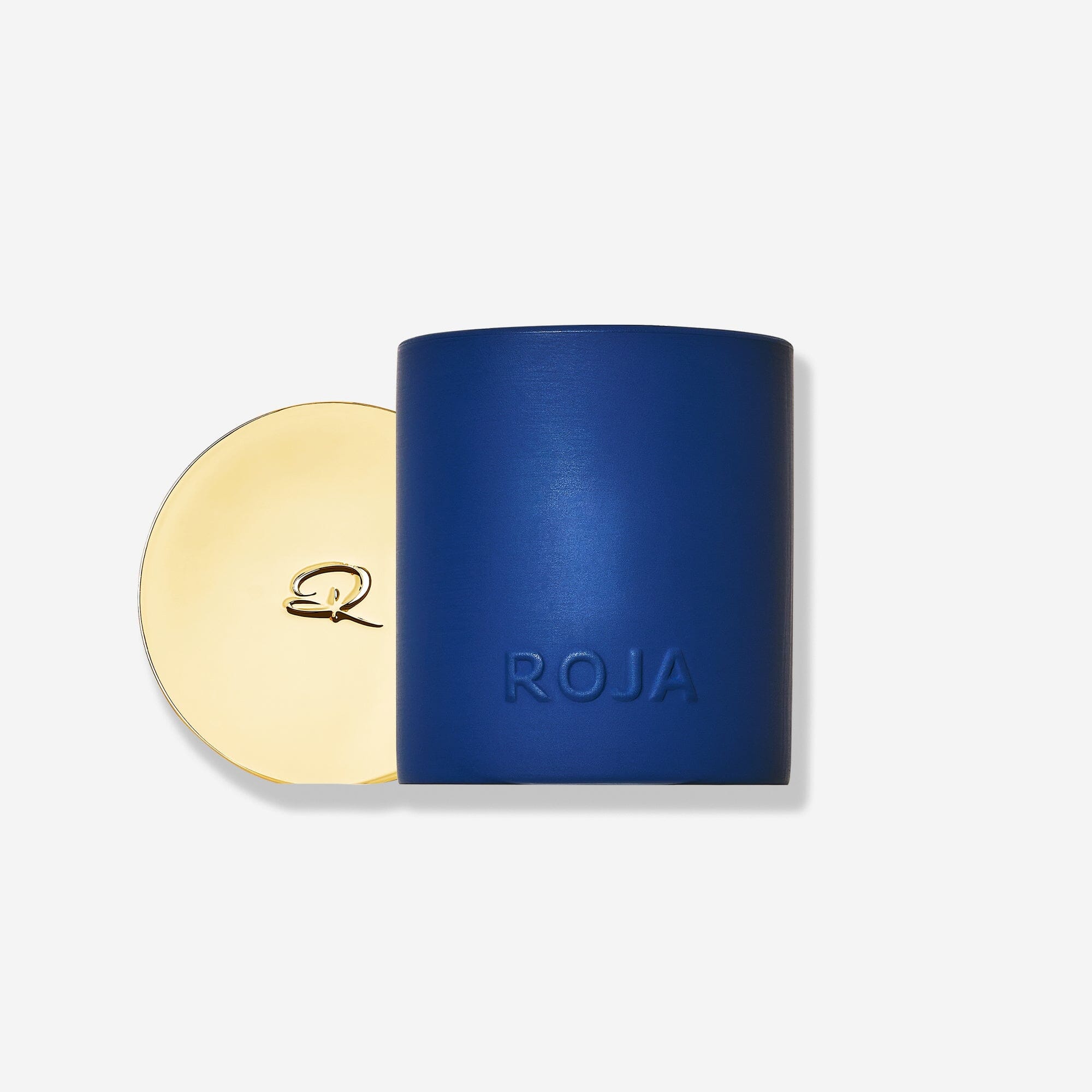 A matte blue cylindrical container embossed with "ROJA" sits beside a round gold lid with a decorative "R," showcasing Roja Parfums’ The Essence Of Christmas candle—a festive home fragrance—set against a plain white background.