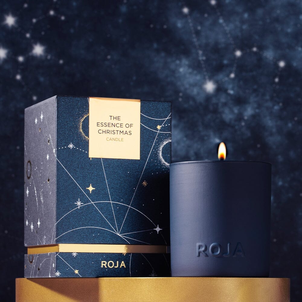 A dark blue candle labeled ROJA sits next to a matching box with gold celestial designs and the label The Essence of Christmas Candle, set against a starry night sky background.