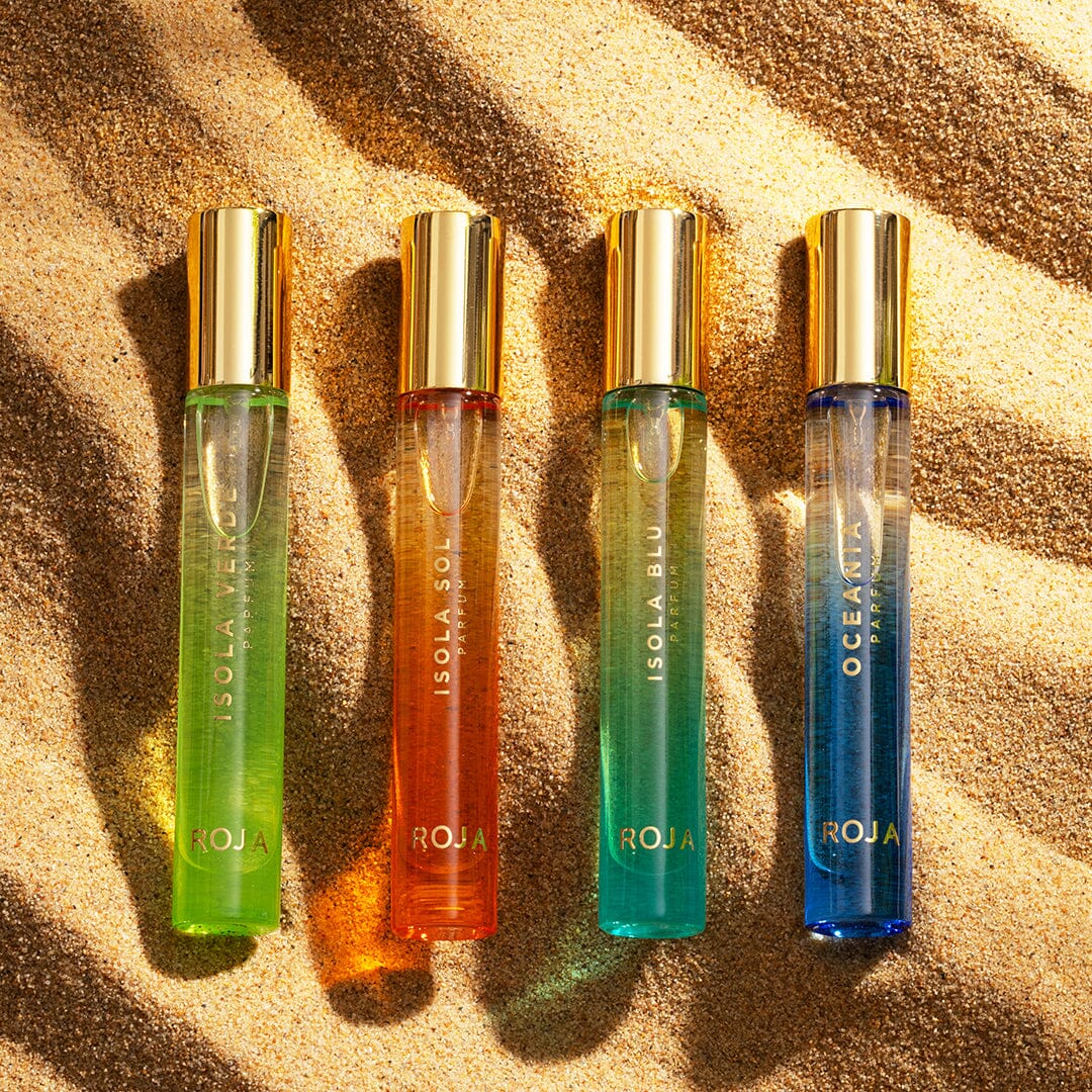 Four vibrant bottles from The Isola Travel Collection by Roja Parfums, each with gold caps, rest on sunlit sand, their shadows and highlights inviting you to experience an evocative sensory journey.