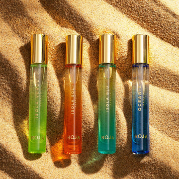 Four vibrant bottles from The Isola Travel Collection by Roja Parfums, each with gold caps, rest on sunlit sand, their shadows and highlights inviting you to experience an evocative sensory journey.