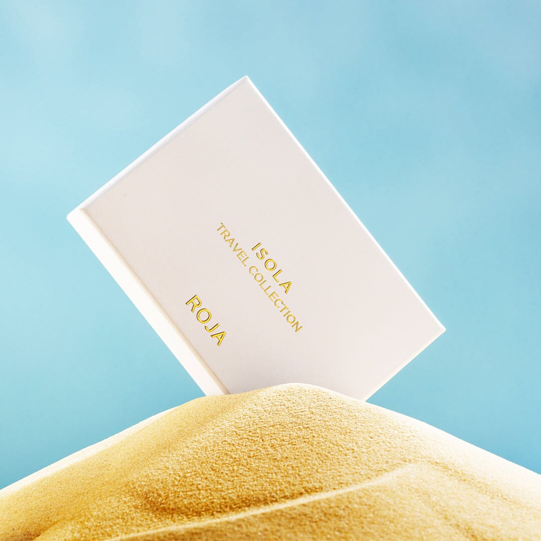 A white box labeled "The Isola Travel Collection" by Roja Parfums rests on yellow sand with a blue backdrop, inviting you to experience an olfactory escape and sensory journey from the Isola Collection.