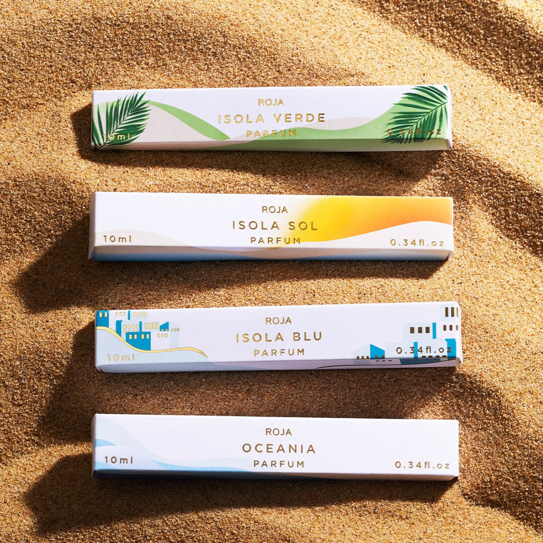 Four rectangular boxes from Roja Parfums Isola Travel Collection—Verde, Sol, Blu, and Oceania—rest on sunlit sand. Each features a colorful, minimalist design reflecting an olfactory escape inspired by its name.