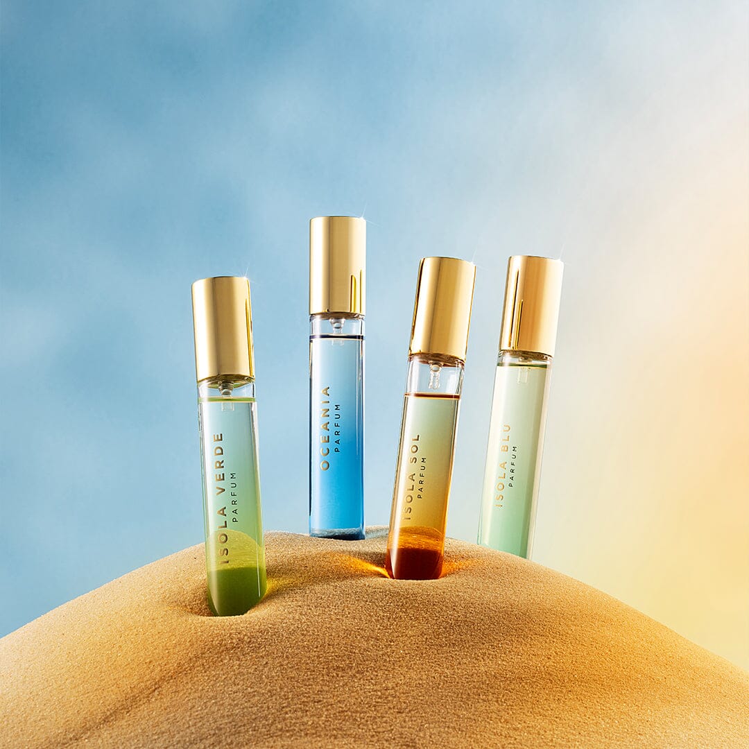 Four bottles from Roja Parfums' The Isola Travel Collection, each with a gold cap, stand upright on sand against a soft sky of blue and gold hues, inviting you to escape through their vibrant elixirs.